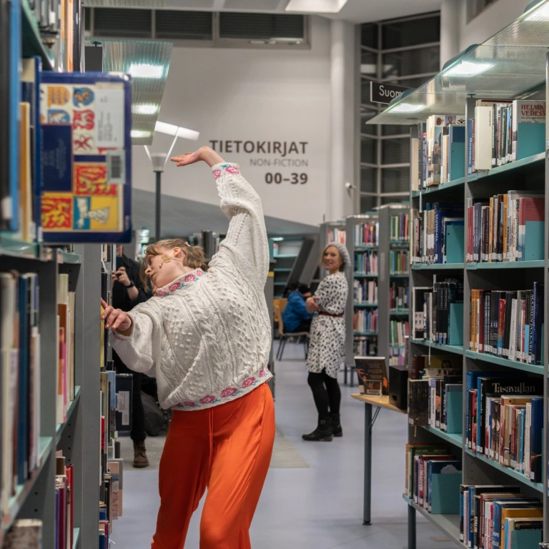 Tanssija liikkuu kirjaston kirjahyllyjen välissä. Taustalla ohikulkija katsoo tanssijaa hymyillen.
