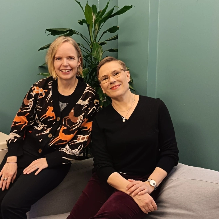 Eeva Eloranta hymyilee Jenni Rissasen vierellä. He istuvat sohvan reunalla takanaan viherkasvi.