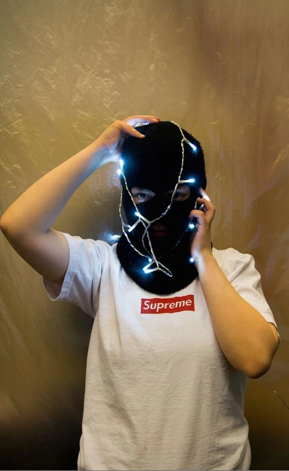 Ihminen valkoisessa paidassa, jossa lukee "Supreme", pitelee käsiään päässään. Hänellä on päässään musta pipo, joka paljastaa vain hänen silmänsä sekä jouluvalonauhaa.
