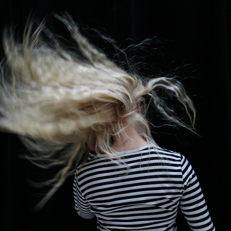 Kiharahiuksinen ihminen heilauttaa hiuksiaan  selkä kohti kameraa. Hänellä on raidallinen paita ja vaaleat hiukset.