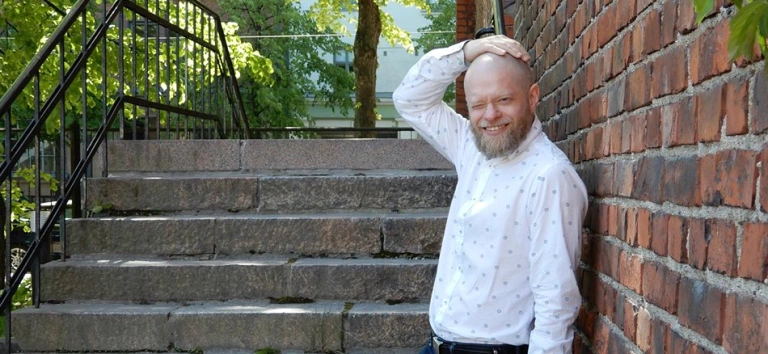 Teppo Virtanen tiiliseinän ja kivisten portaiden vieressä kesäisenä päivänä. Hän hymyilee valkoisessa kauluspaidassa ja pitelee oikeaa kättään päänsä päällä..