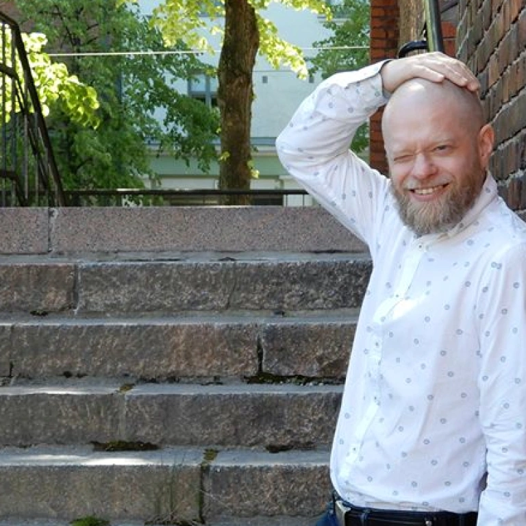 Teppo Virtanen tiiliseinän ja kivisten portaiden vieressä kesäisenä päivänä. Hän hymyilee valkoisessa kauluspaidassa ja pitelee oikeaa kättään päänsä päällä..