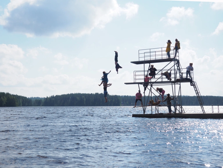 Joukko ihmisiä hyppytornissa järvellä. Muutama hyppää tornista veteen ja loput katsovat tornin eri tasoilla. Aurinkoinen päivä.