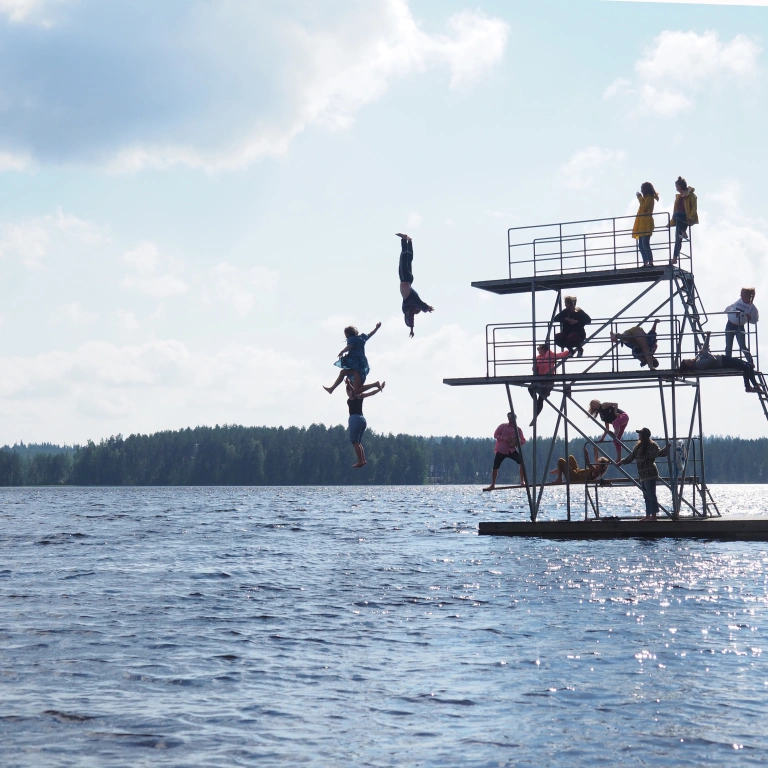 Joukko ihmisiä hyppytornissa järvellä. Muutama hyppää tornista veteen ja loput katsovat tornin eri tasoilla. Aurinkoinen päivä.