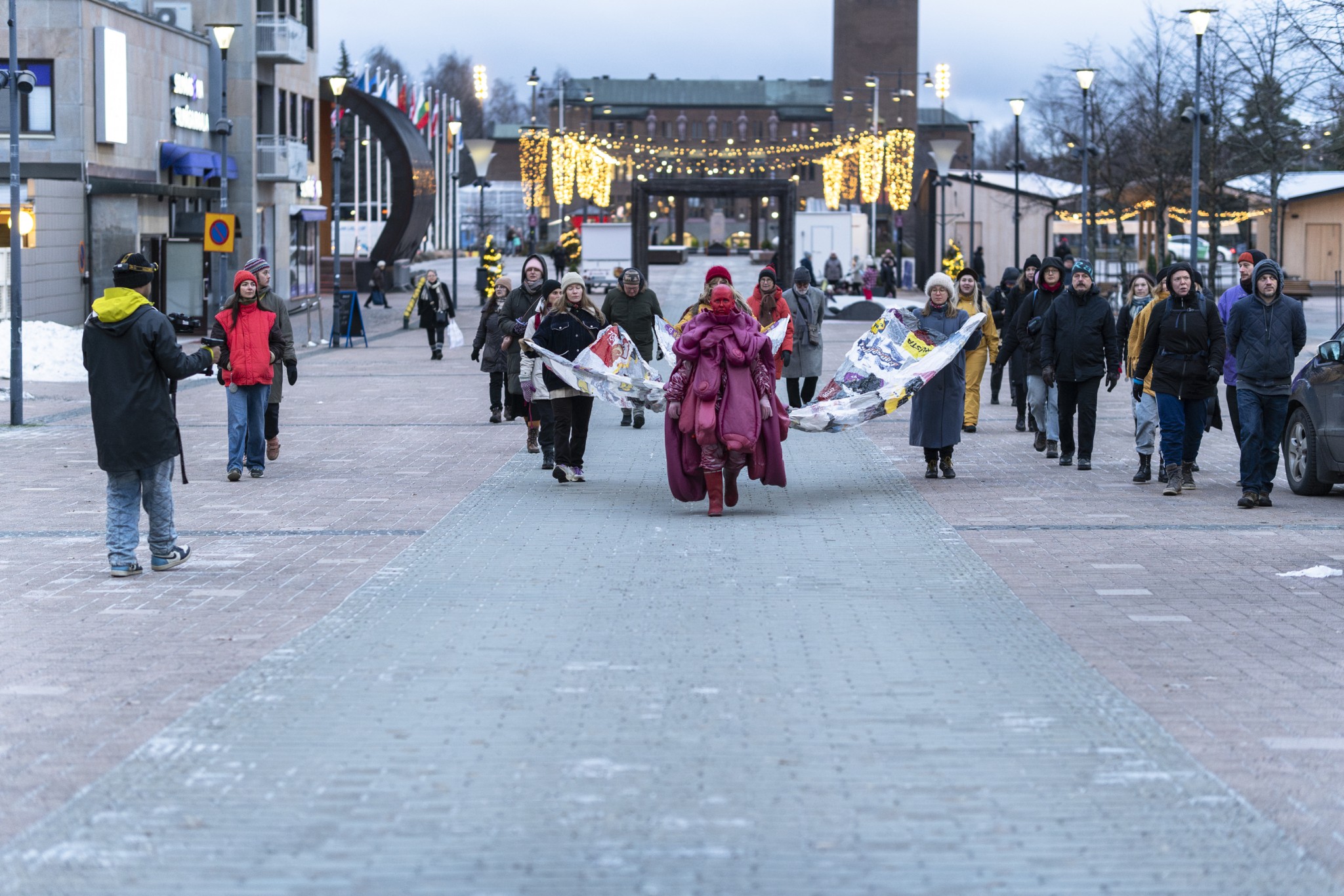 Joukko ihmisiä kulkee Joensuun torilla harmaassa talvisäässä. Joukkoa johtaa punaiseen näyttävään asuun pukeutunut, punaiseksi maalattu esiintyjähahmo.