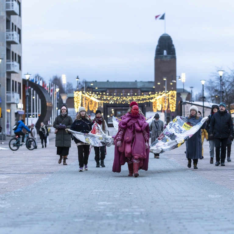Punaiseen näyttävään suomukkaaseen pukuun pukeutunut ja punaiseksi maalattu esiintyjä johtaa joukkoa, jotka pitelevät hänen puvustaan lähtevää muovipusseista ommeltua laahusta. Taustalla Joensuun kaupunginteatterin torni ja harmaa taivas Joensuun torin yllä.